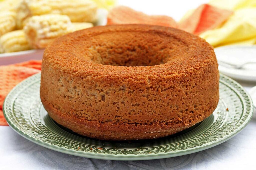 Bolo redondo com furo no meio sobre prato que tem detalhes na borda, ao fundo panos e espigas de milho em uma travessa