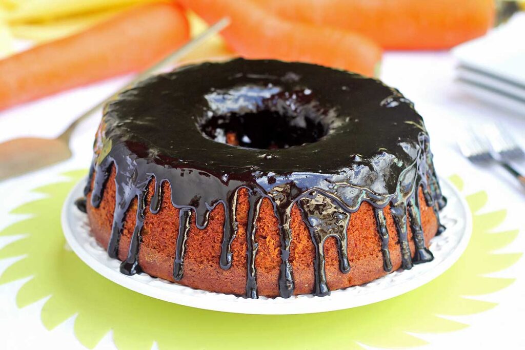 Bolo de cenoura redondo com furo no meio, sobre o bolo uma cobertura brilhante de chocolate, ao fundo algumas cenouras