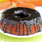 Bolo de cenoura redondo com furo no meio, sobre o bolo uma cobertura brilhante de chocolate, ao fundo algumas cenouras