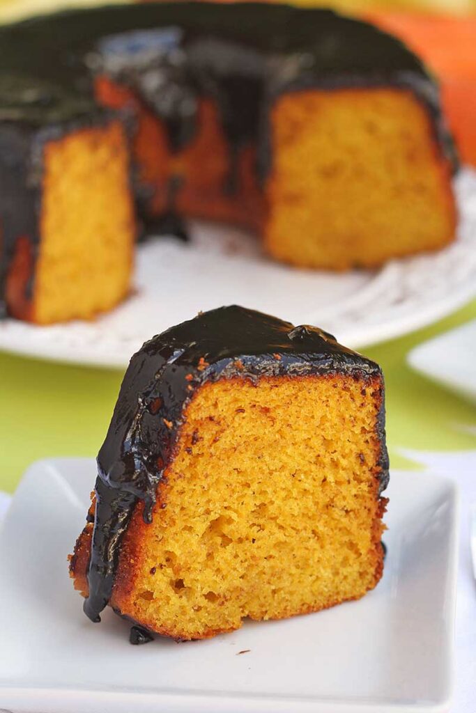 fatia de bolo de cenoura com cobertura de chocolate sobre prato quadrado pequeno, ao fundo o bolo partido desfocado