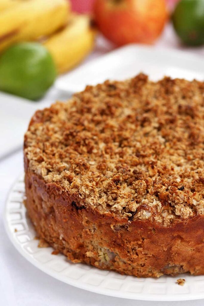 Torta redonda com farofa crocante por cima, banana, maçã e limão ao fundo desfocados