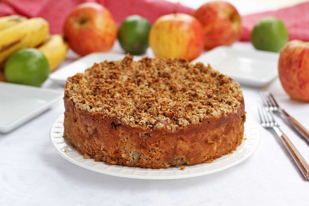 Torta redonda com farofa crocante por cima, ao fundo bananas, maçãs e alguns limões ao lado de alguns pratos quadrados pequenos