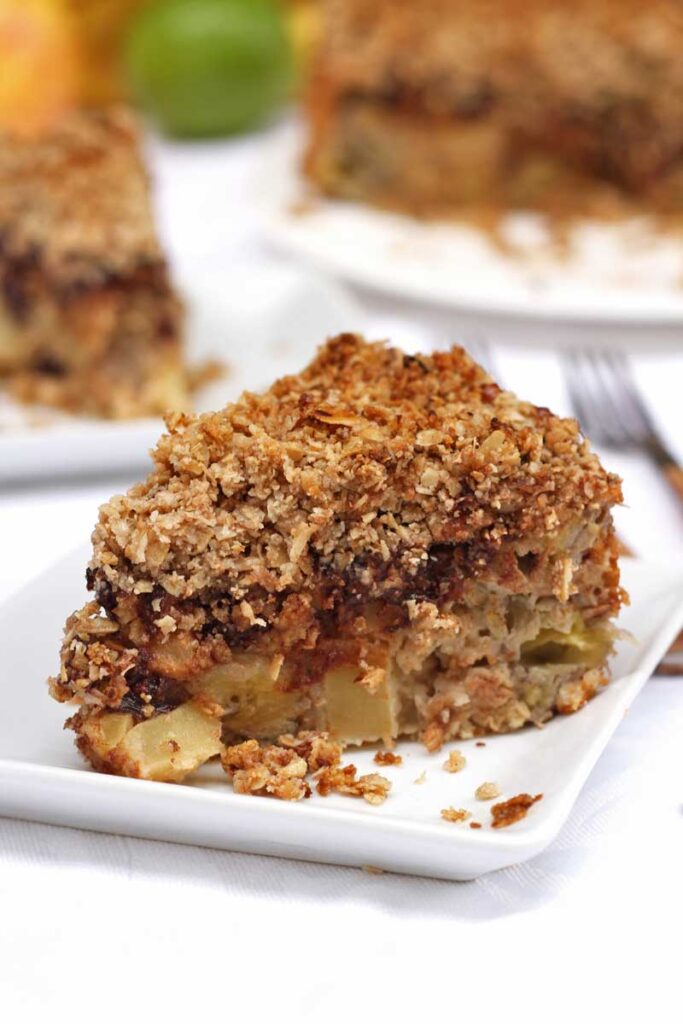 Fatia de torta de banana e maçã, sobre as frutas tem um pouco de chocolate em pedaços e por cima uma farofinha crocante de flocos de aveia com farinha de amêndoas