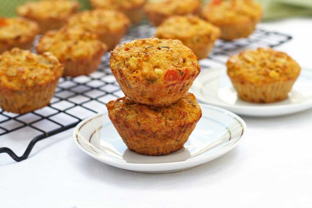 Dois muffins salgados sobrepostos sobre prato redondo pequeno, ao fundo mais muffins espalhados sobre uma grade
