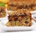 Fatia de torta de banana e maçã com cobertura crocante de aveia em flocos finos e farinha de amêndoas, no meio da torta tem uva passa e sobre as frutas tem pedaços de chocolate, na sequência vem a farofa