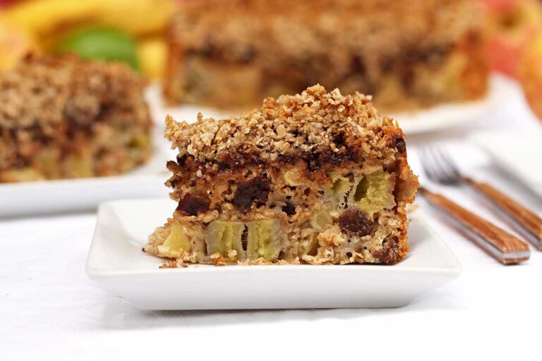 TORTA DE BANANA E MAÇÃ SEM AÇÚCAR SEM TRIGO SEM LEITE Fatia de torta de banana e maçã com cobertura crocante de aveia em flocos finos e farinha de amêndoas, no meio da torta tem uva passa e sobre as frutas tem pedaços de chocolate, na sequência vem a farofa