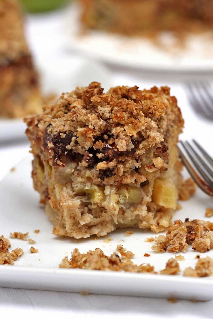 Fatia de torta de banana e maçã com chocolate e cobertura crocante, farelos da cobertura ao lado da fatia e um garfinho pequeno ao lado, a fatia já foi comida alguns pedaços