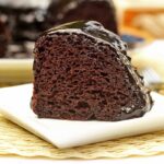 Fatia de bolo de chocolate com cobertura brilhante e cremosa por cima, o bolo está sobre prato quadrado pequeno. Ao fundo o bolo que já foi partido