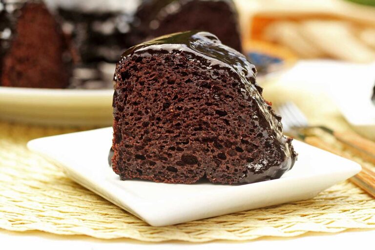 Fatia de bolo de chocolate com cobertura brilhante e cremosa por cima, o bolo está sobre prato quadrado pequeno. Ao fundo o bolo que já foi partido