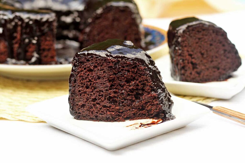 duas fatias de bolo de chocolate sobre dois pratos quadrados pequenos, a fatia da frente já foi parcialmente comida, já tem uma garfada a menos na fatia, ao fundo o bolo partido