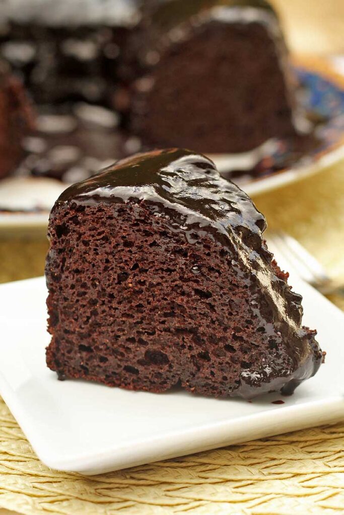 Fatia de bolo de chocolate com cobertura cremosa de chocolate sobre prato quadrado pequeno, oa fundo o bolo partido