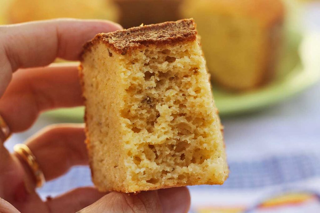 Mão segurando fatia de bolo de fubá já mordida, mostrando textura aerada da massa