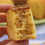 Mão segurando fatia de bolo de fubá já mordida, mostrando textura aerada da massa