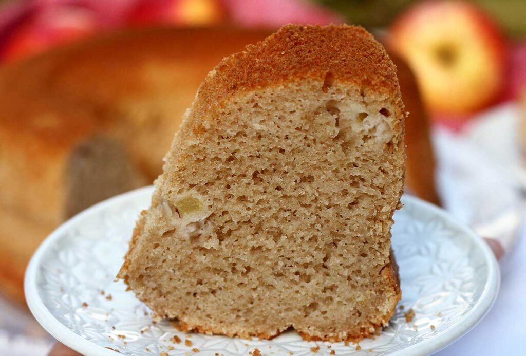Fatia de bolo de maçã sobre prato redondo pequeno, a foto está bem aproximada da fatia, mostrando o bolo partido de fundo e umas maçãs desfocadas