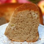 Fatia de bolo de maçã sobre prato redondo pequeno, a foto está bem aproximada da fatia, mostrando o bolo partido de fundo e umas maçãs desfocadas