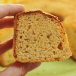 Mão segurando fatia de bolo de fubá, ao fundo o bolo partido desfocado