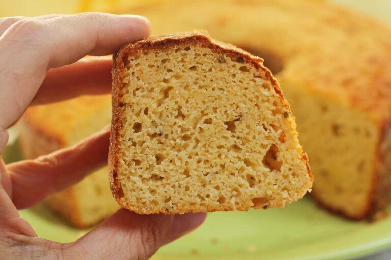 BOLO DE FUBÁ COM ERVA-DOCE SEM AÇÚCAR Mão segurando fatia de bolo de fubá, ao fundo o bolo partido desfocado