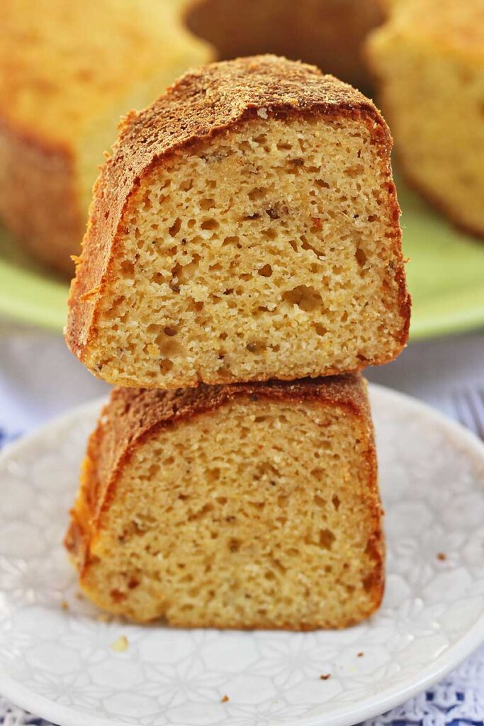 Duas fatias de bolo de fubá sobrepostas em um prato redondo pequeno, ao fundo o bolo partido desfocado