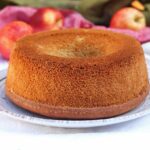 Bolo redondo com furo no meio sobre prato branco, ao fundo várias maçãs e um pano verde e vermelho