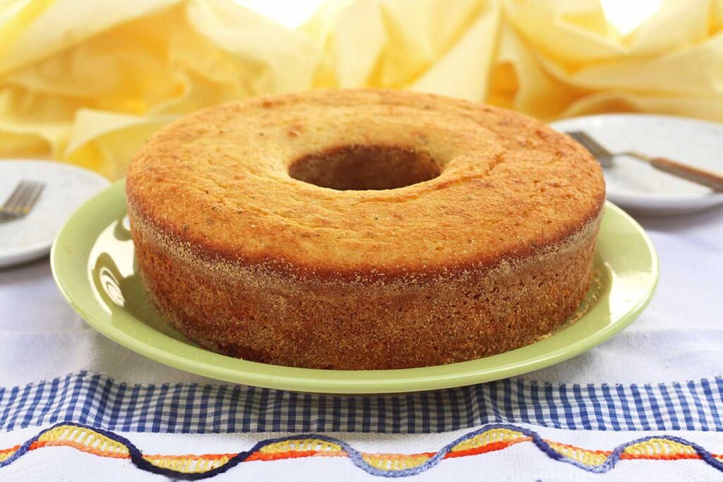 Bolo de fubá simples, redondo com furo no meio, embaixo do prato um pano de louça e ao fundo um pano amarelo combinando com o bolo