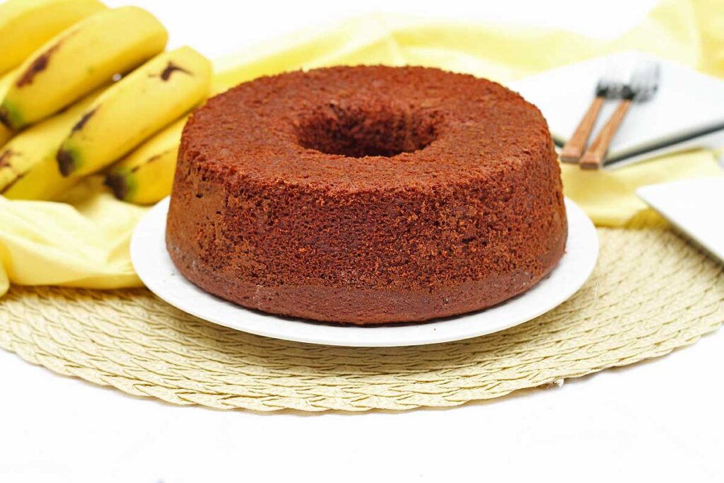 Bolo redondo com furo no meio ao fundo um cacho de banana e pratinhos quadrados pequenos