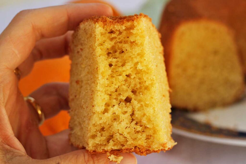 Mão segurando fatia de bolo de laranja já mordido, mostrando a textura aerada da massa