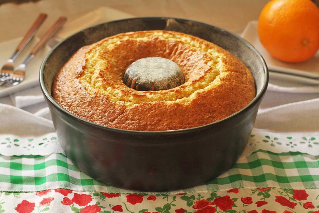 Bolo de laranja dentro de uma forma redonda com furo no meio, o bolo está no topo da forma, bem douradinho, ao fundo alguns pratinhos e garfos e uma laranja