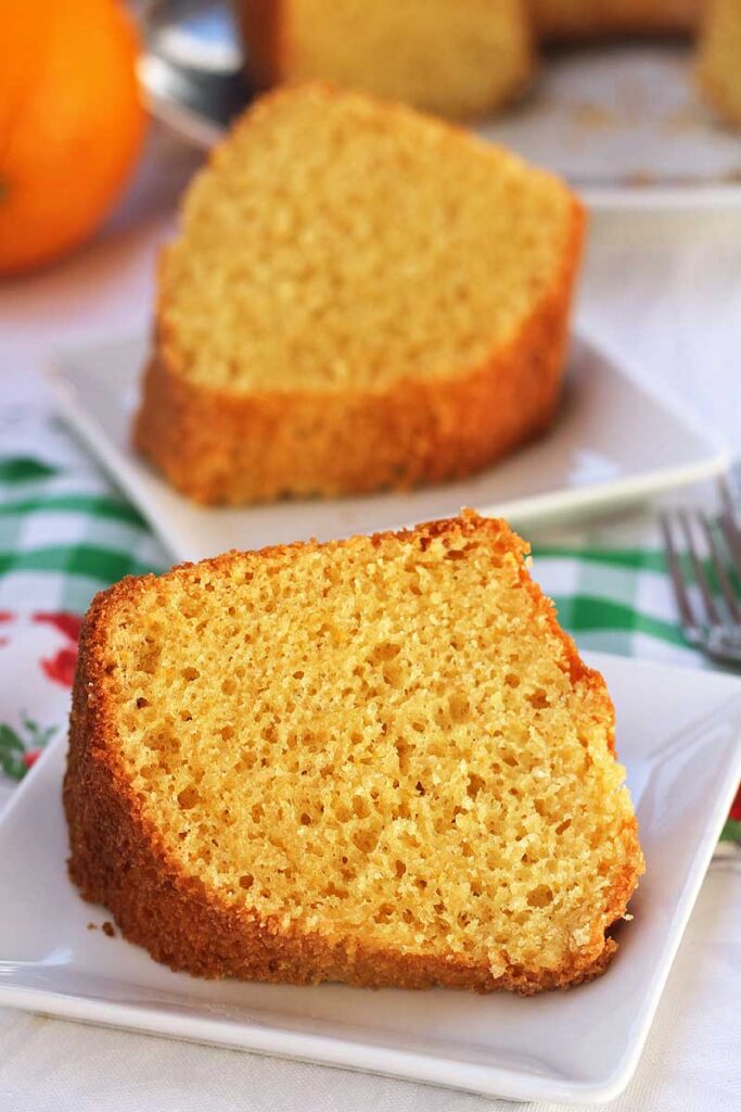 Duas fatias de bolo de laranja, cada uma está em uma das fatias estão deitadas sobre um prato quadrado pequeno, os pratos estão sobre um pano de prato