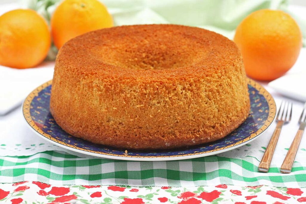 bolo redondo com furo no meio sobre prato redondo, o prato está sobre um pano de prato branco com detalhez xadrez verde e flores vermelhas, ao fundo três laranjas