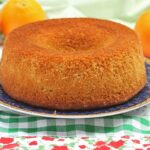 bolo redondo com furo no meio sobre prato redondo, o prato está sobre um pano de prato branco com detalhez xadrez verde e flores vermelhas, ao fundo três laranjas