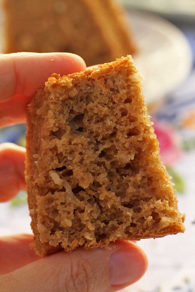 mão segurando pedaço de bolo de coco mordido, mostrando a textura bem aerada 