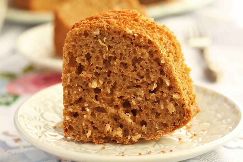 fatia de bolo de coco sobre prato redondo pequeno, na fatia da pra perceber os flocos de coco que são moídos grossos