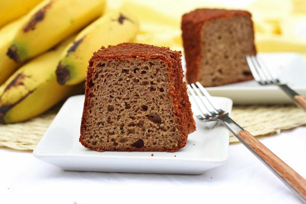 Duas fatias sobre 2 pratos quadrados pequenos, ao fundo um cacho de banana, ao lado de cada prato, um garfinho