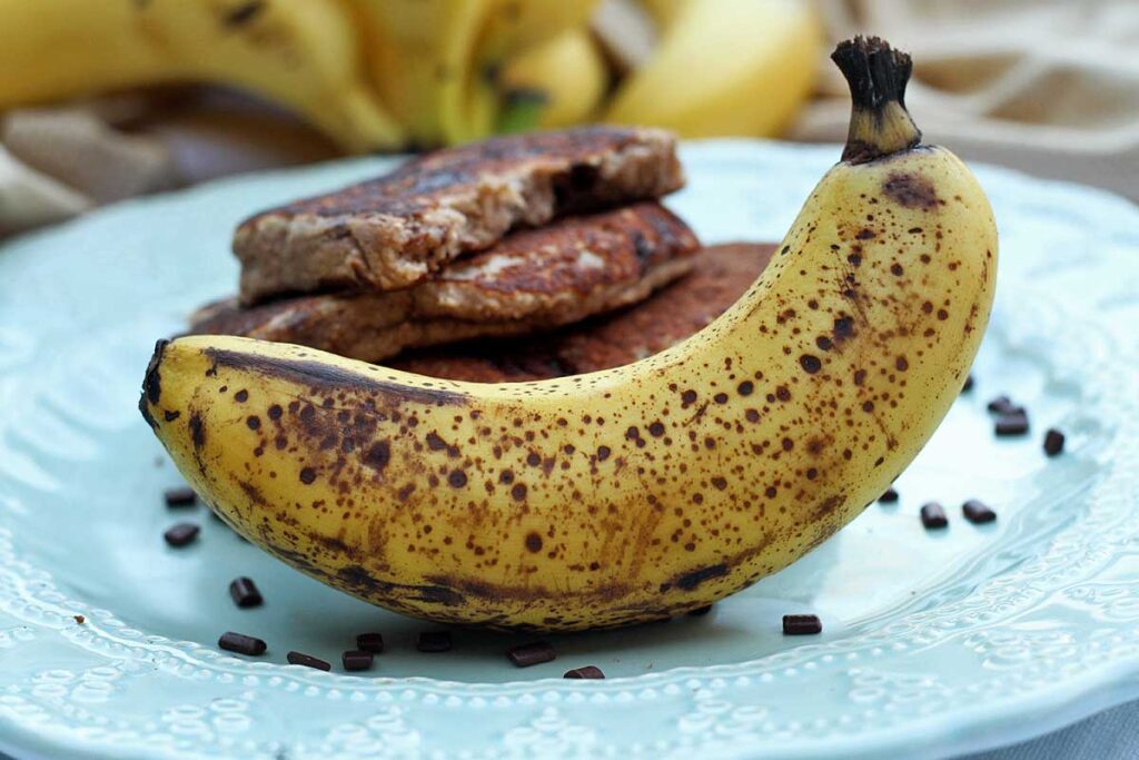Banana madura com pontos pretos na casca, sobre prato que tem as panquecas ao fundo