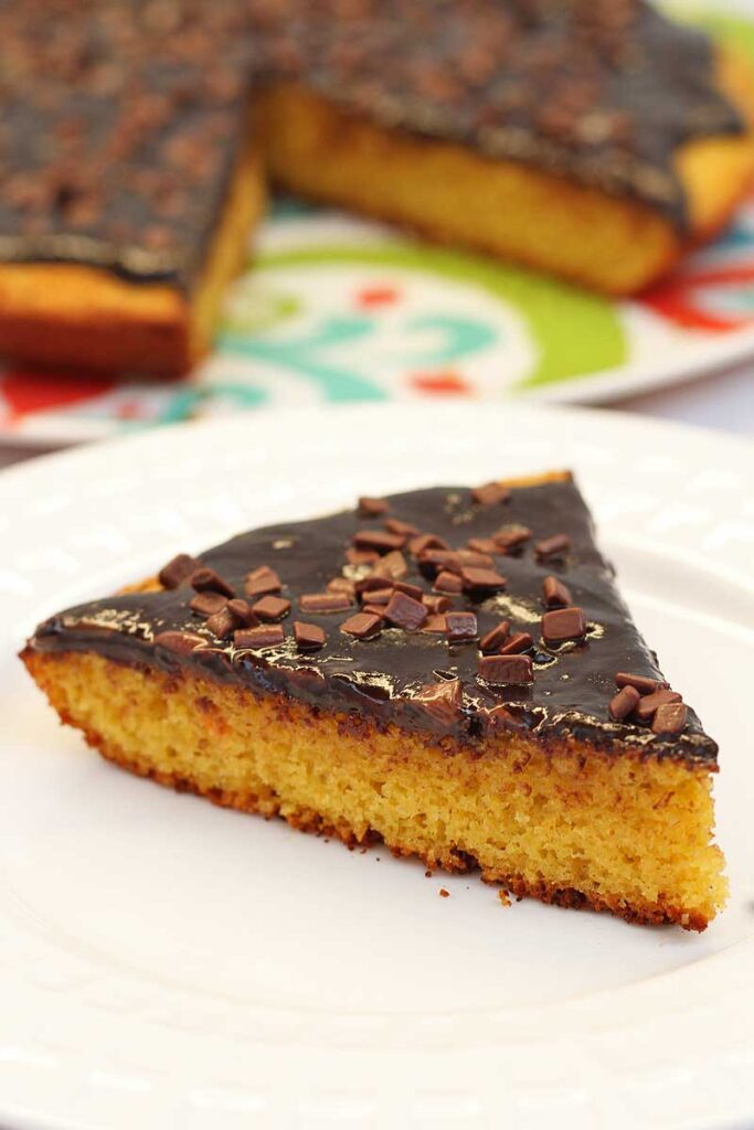 fatia triangular de bolo de cenoura com cobertura de chocolate, no topo da cobertura granulado de chocolate e ao fundo o bolo redondo faltando um pedaço