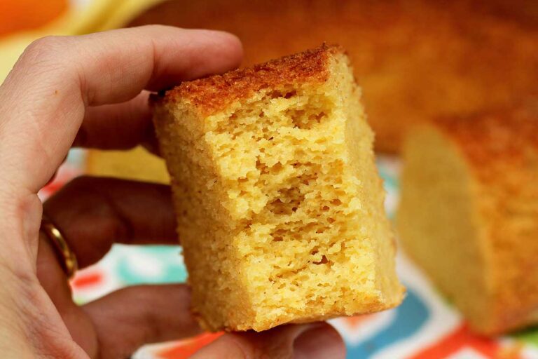 BOLO DE FUBÁ COM LARANJA E AVEIA DE LIQUIDIFICADOR mão segurando fatia de bolo já mordida, mostrando textura bem aerada, ao fundo o bolo partido e desfocado