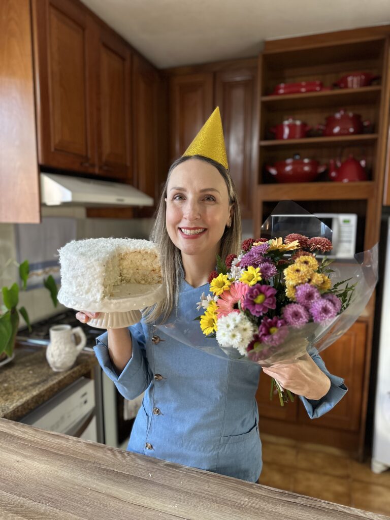 mulher loira segurando bolo já cortado em uma mão e um buquê de flores na outra mão, ela está usando uniforme de chef de cozinha e um chapeuzinhi de aniversário