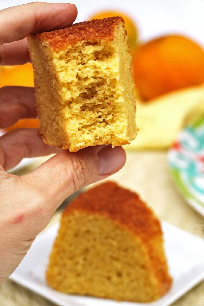 Mão segurando fatia de bolo de fubá com laranja já mordido, mostrando a textura bem aerada e leve da massa, abaixo uma fatia inteira de bolo sobre um prato quadrado pequeno