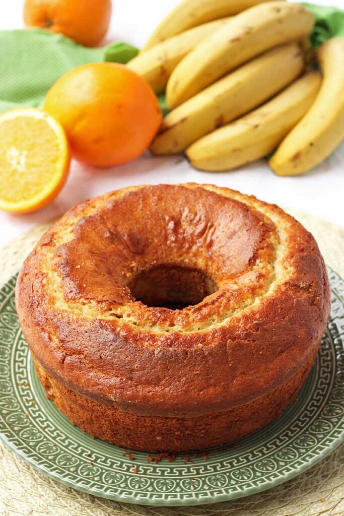 Bolo redondo com furo no meio, ao fundo uma penca de banana, uma laranja inteira e uma partida ao meio