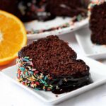 Duas fatias de bolo de chocolate com cobertura de chocolate, cada uma em um prato quadrado pequeno, a da frente está deitada e a de trás está de pé, tem uma laranja cortada ao meio ao lado do prato que está na frente