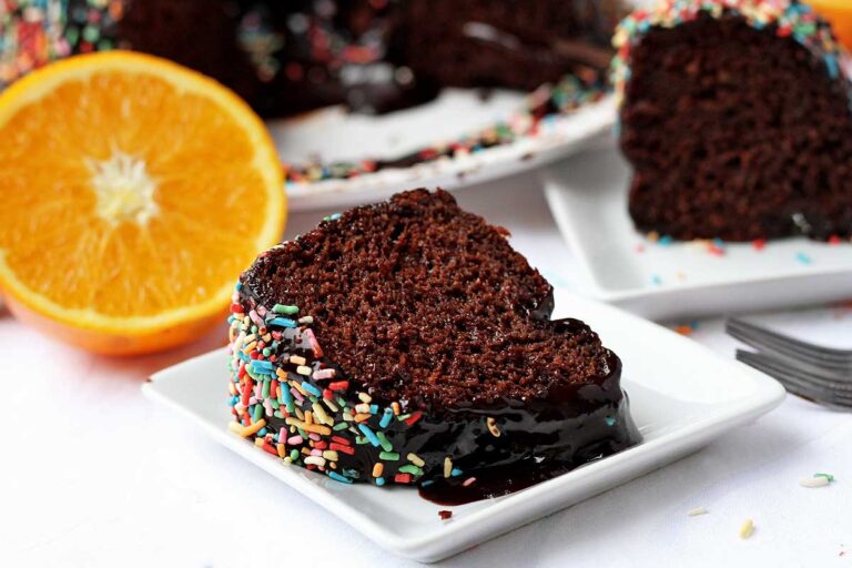 Duas fatias de bolo de chocolate com cobertura de chocolate, cada uma em um prato quadrado pequeno, a da frente está deitada e a de trás está de pé, tem uma laranja cortada ao meio ao lado do prato que está na frente