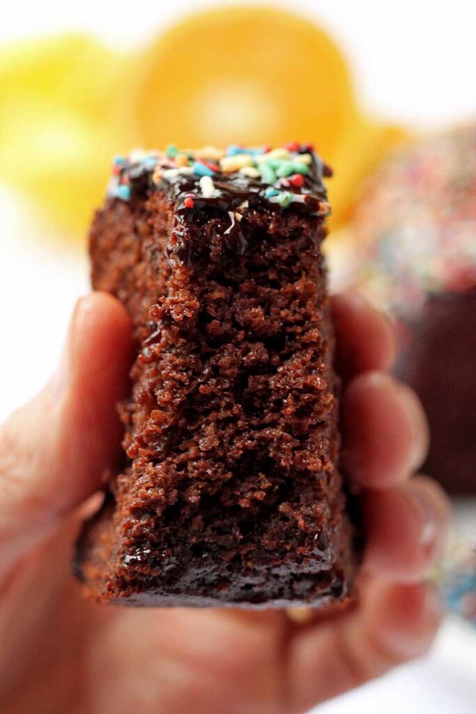 Mão segurando fatia de bolo de chocolate com cobertura de chocolate e granulado colorido, a fatia já está mordida, mostrando a textura aerada do bolo