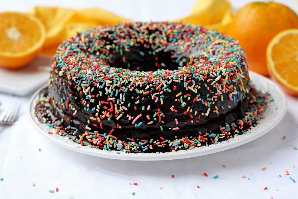 bolo redondo com furo no meio com chocolate, cobertura de chocolate e granulado colorido por cima, ao fundo laranjas
