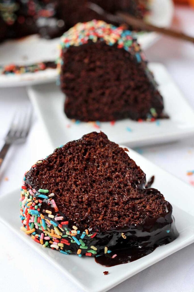 Duas fatias de bolo de chocolate com cobertura de chocolate, cada uma em um prato quadrado pequeno, a da frente está deitada e a de trás está de pé