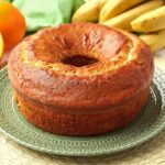 Bolo redondo com furo no meio, ao fundo uma penca de banana, uma laranja inteira e uma partida ao meio