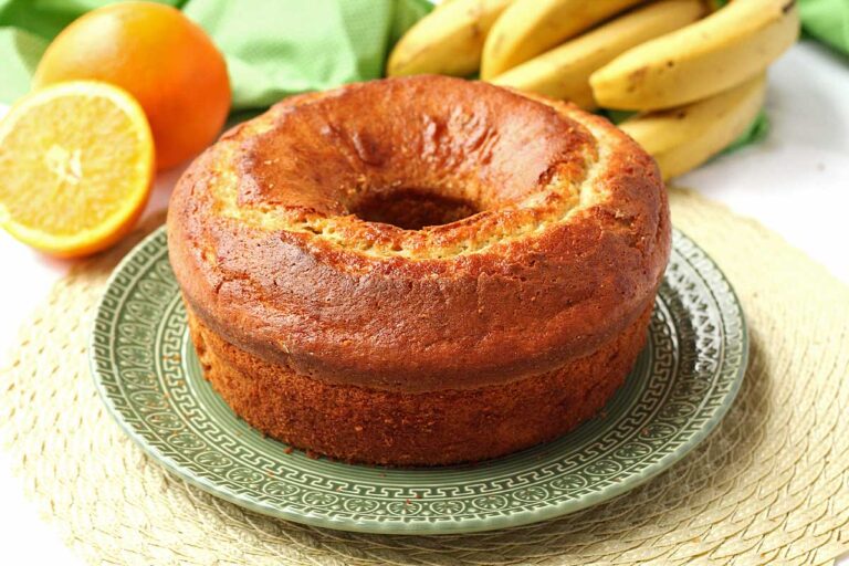BOLO DE BANANA E LARANJA DE LIQUIDIFICADOR Bolo redondo com furo no meio, ao fundo uma penca de banana, uma laranja inteira e uma partida ao meio