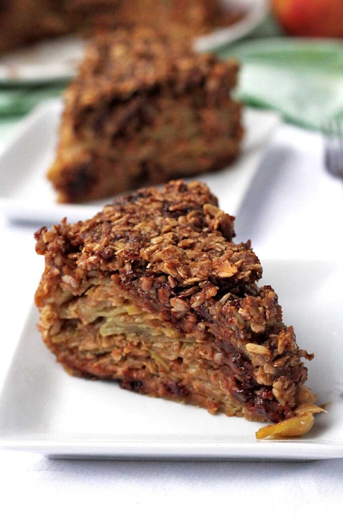 Duas fatias triangulares de tprta de maçã com cobertura de aveia e pedaços de chocolate entre as fatias de maçã, cada fatia está sobre um prato quadrado pequeno