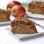 Duas fatias triangulares de torta de maçã com cobertura de aveia com açúcar mascavo, ao lado do pratinho 2 garfos e ao fundo 2 maçãs