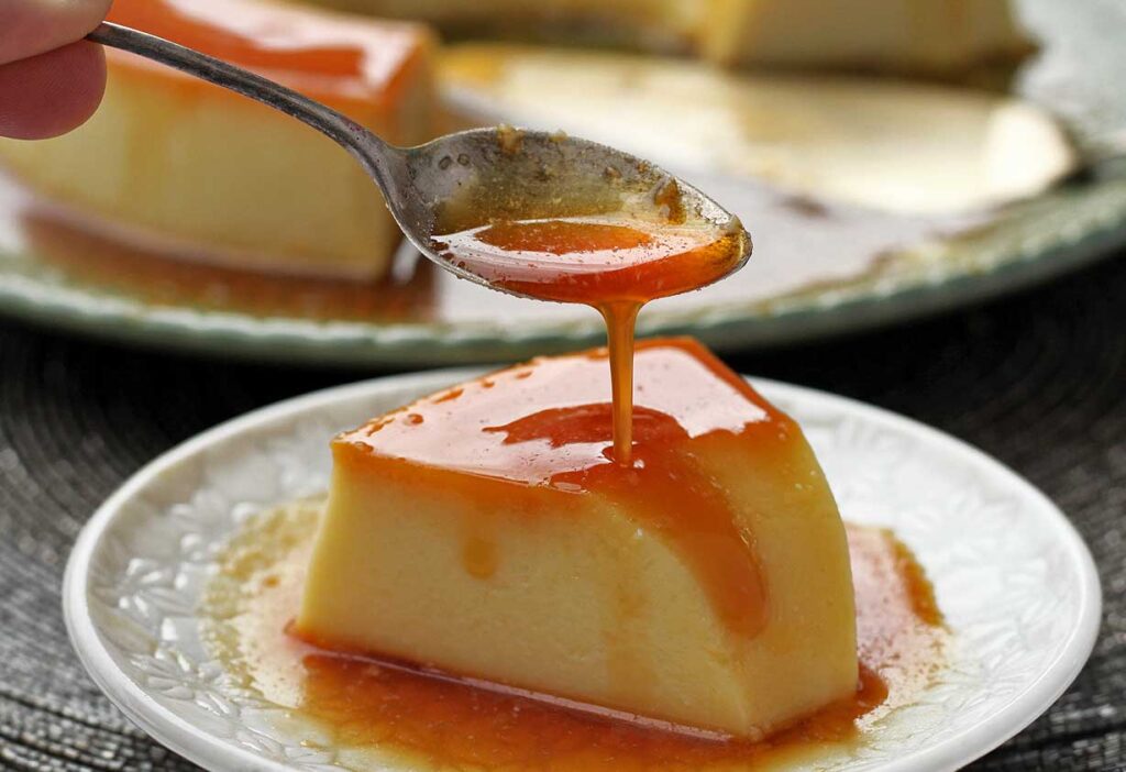 fatia de pudim dobre prato redondo pequeno, acima da fatia uma colher pequena derramando caramelo por cima