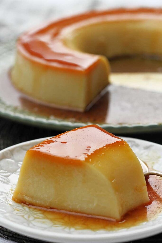 fatia de pudim de caramelo sobre prato redondo pequeno, ao fundo o pudim já cortado